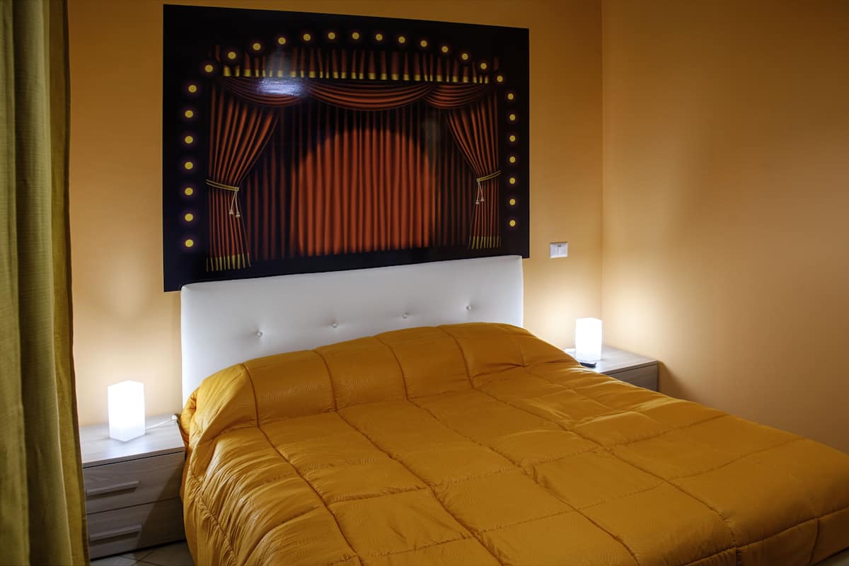 A bed with a headboard featuring a curtain design and lights, a bedside table with a lamp, and a curtain partially visible on the left.