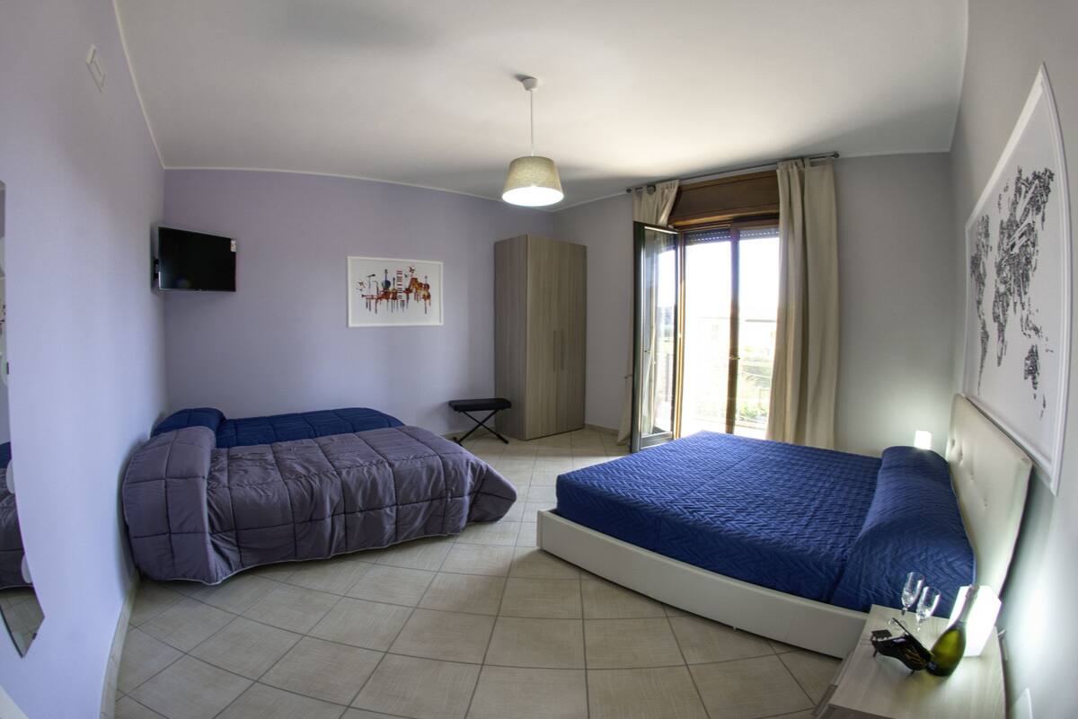 A bedroom with two beds, a TV, a window with curtains, and a small table with a bottle.