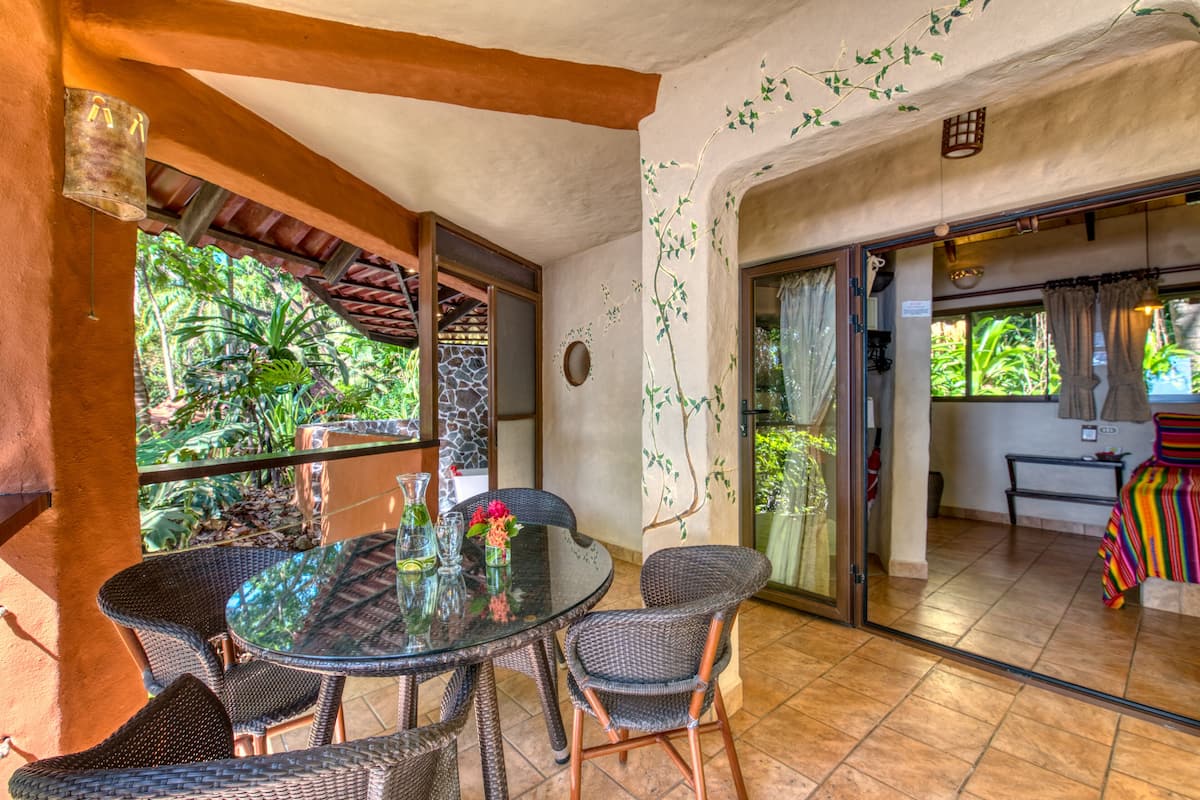 A dining area with a glass table and wicker chairs, a stone wall, and a view of greenery outside.