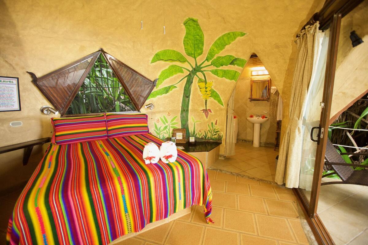A bed with a colorful striped bedspread and two pillows, a wall with a palm tree mural, a bathroom with a sink and mirror, and a staircase leading to an outdoor area.