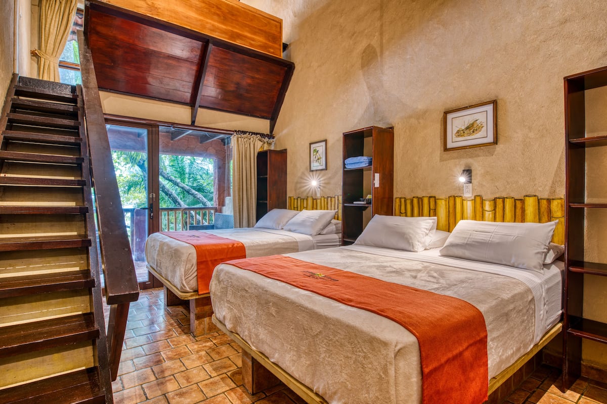 A room with two beds, wooden furniture, a staircase, and a view of the outdoors.