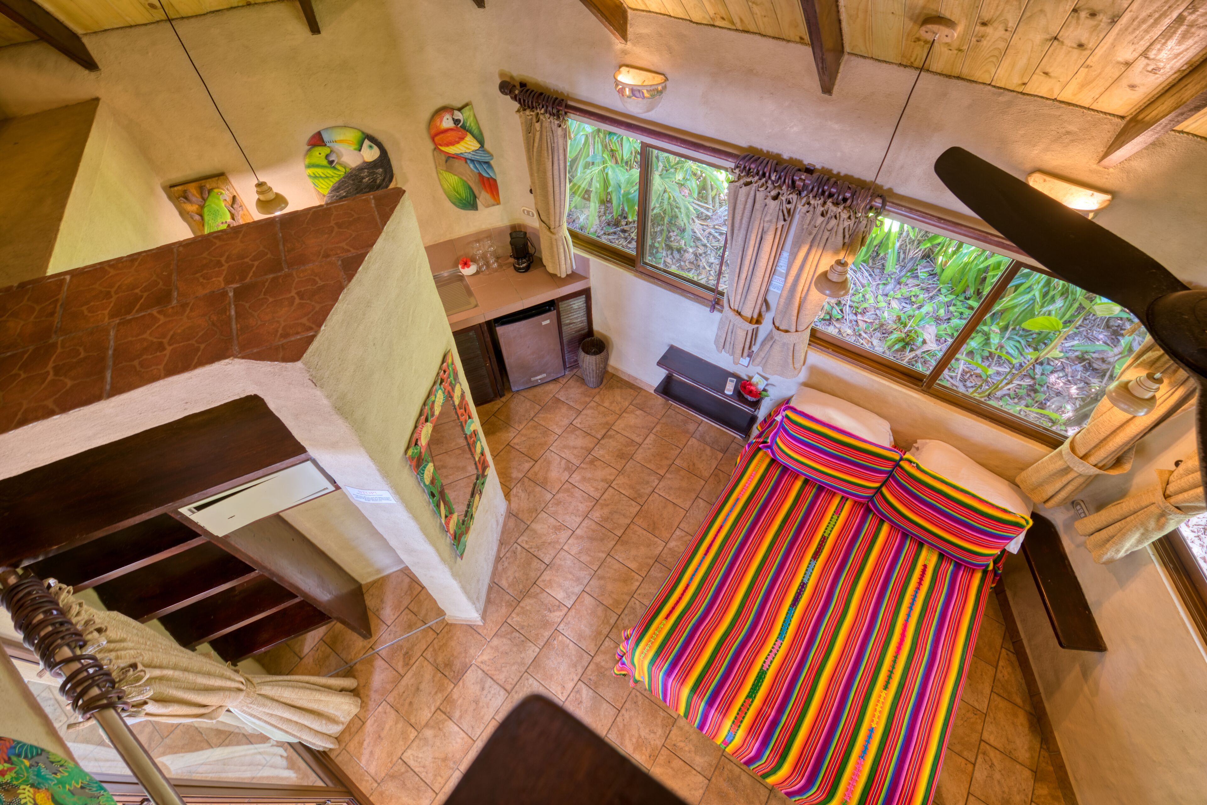 A room with a bed, a small kitchenette, a window with curtains, and a view of greenery outside.