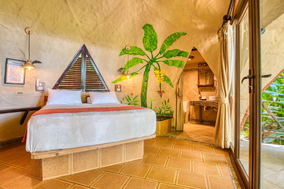 A bedroom with a bed, a wall-mounted lamp, a framed travel poster, and a potted plant.