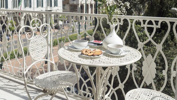Un balcón con mobiliario de hierro forjado blanco, una mesa con una taza, un platillo y un florero con una sola rosa amarilla.