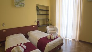 Habitación de hotel con dos camas, una silla, una estantería con una planta y una ventana con cortinas.