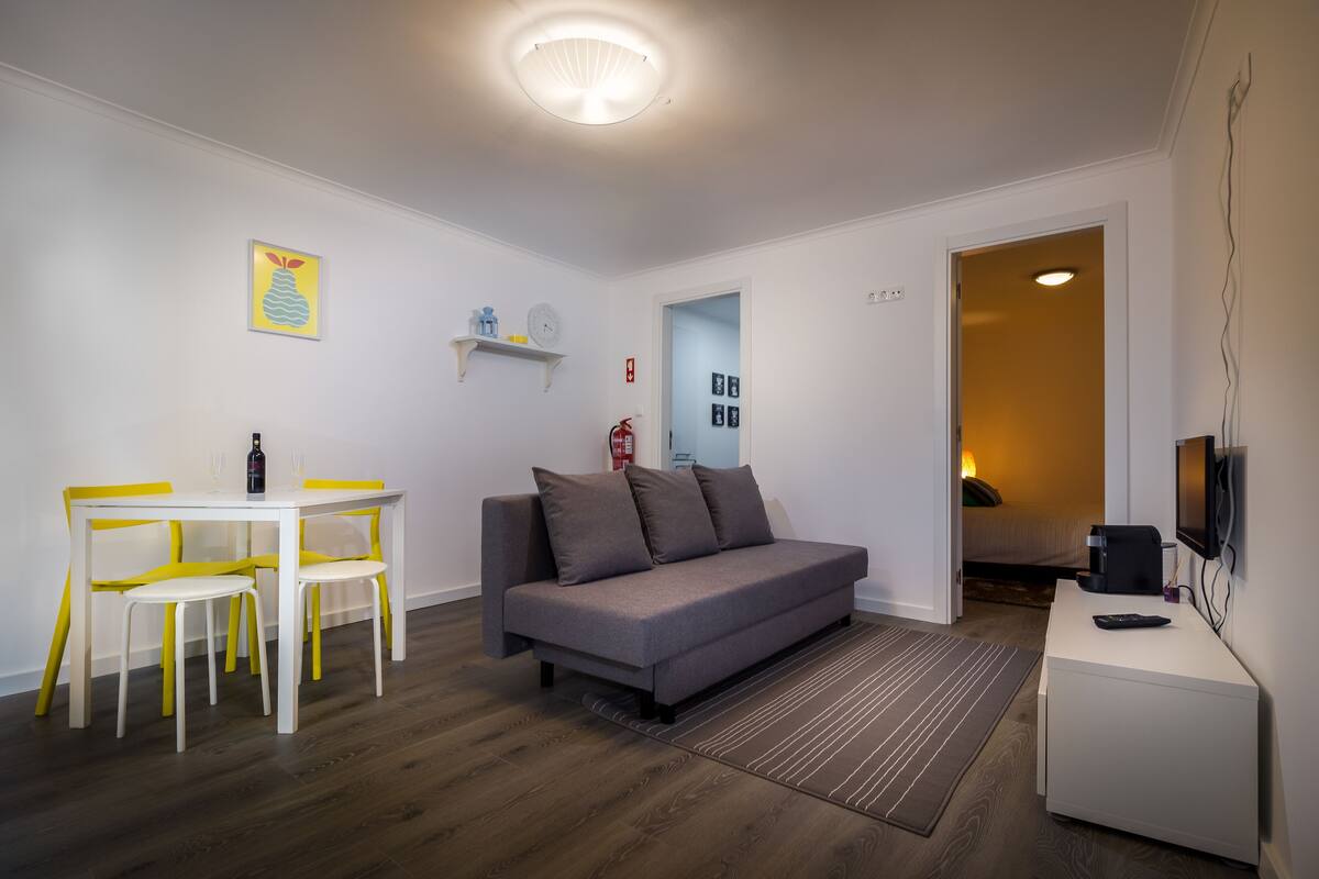Uma sala de estar moderna com um sofá cinzento, poltronas amarelas, uma mesa branca e uma televisão de tela plana.