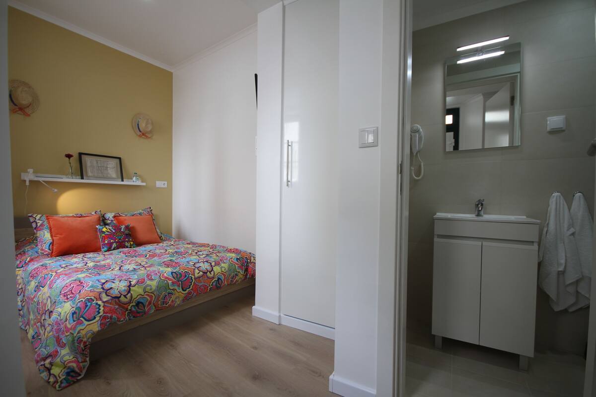 Quarto com colcha colorida, guarda-roupa branco e banheiro com pia e espelho.