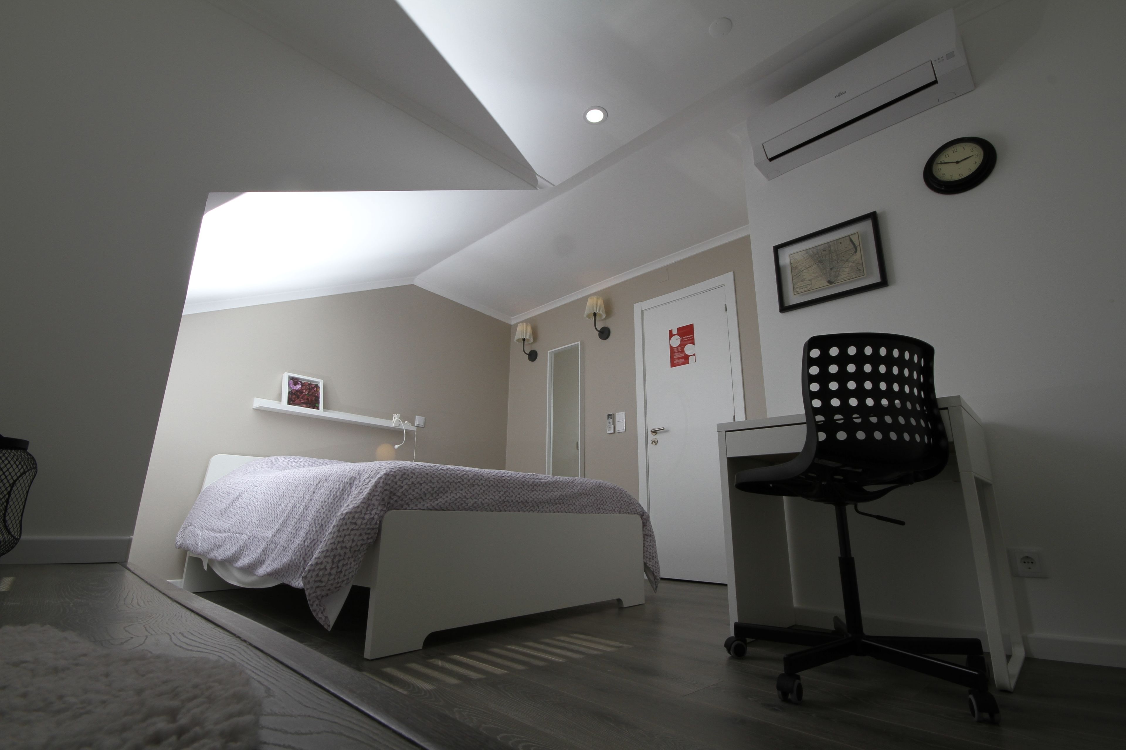 A bedroom with a bed, a desk, a chair, an air conditioner, a clock, and a door.