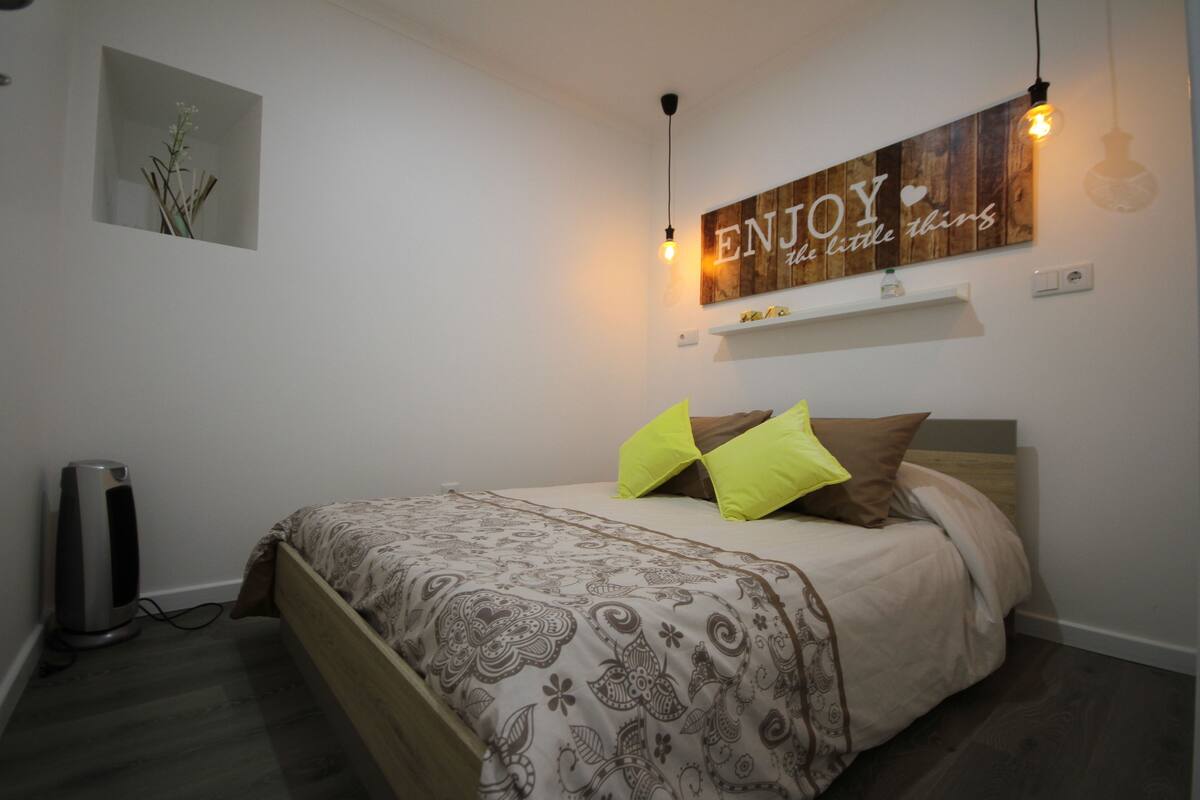 Uma cama bem arrumada com roupa de cama estampada, uma prateleira na parede, uma placa decorativa e um aparelho de luz fixado na parede.