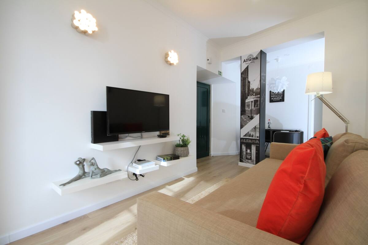 Uma sala de estar moderna com uma TV de tela plana fixada em uma prateleira branca, um sofá bege com um travesseiro vermelho e um abajur.