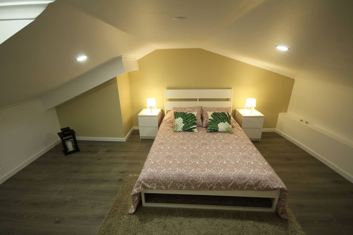 Quarto com teto inclinado, cama com roupa de cama estampada, duas luminárias de cabeceira e um pequeno candeeiro preto no chão.