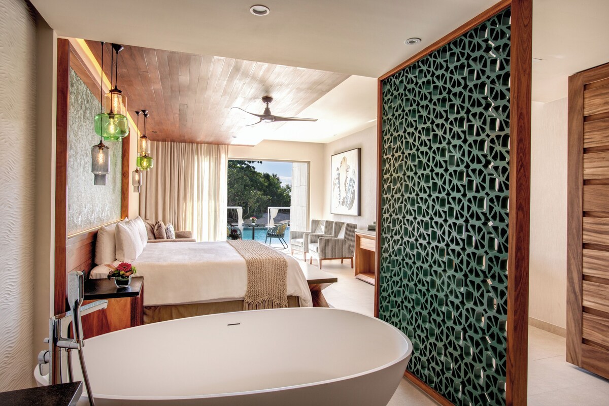 Habitación de hotel moderna con una bañera grande, una cama, un área de comedor y vistas al exterior.