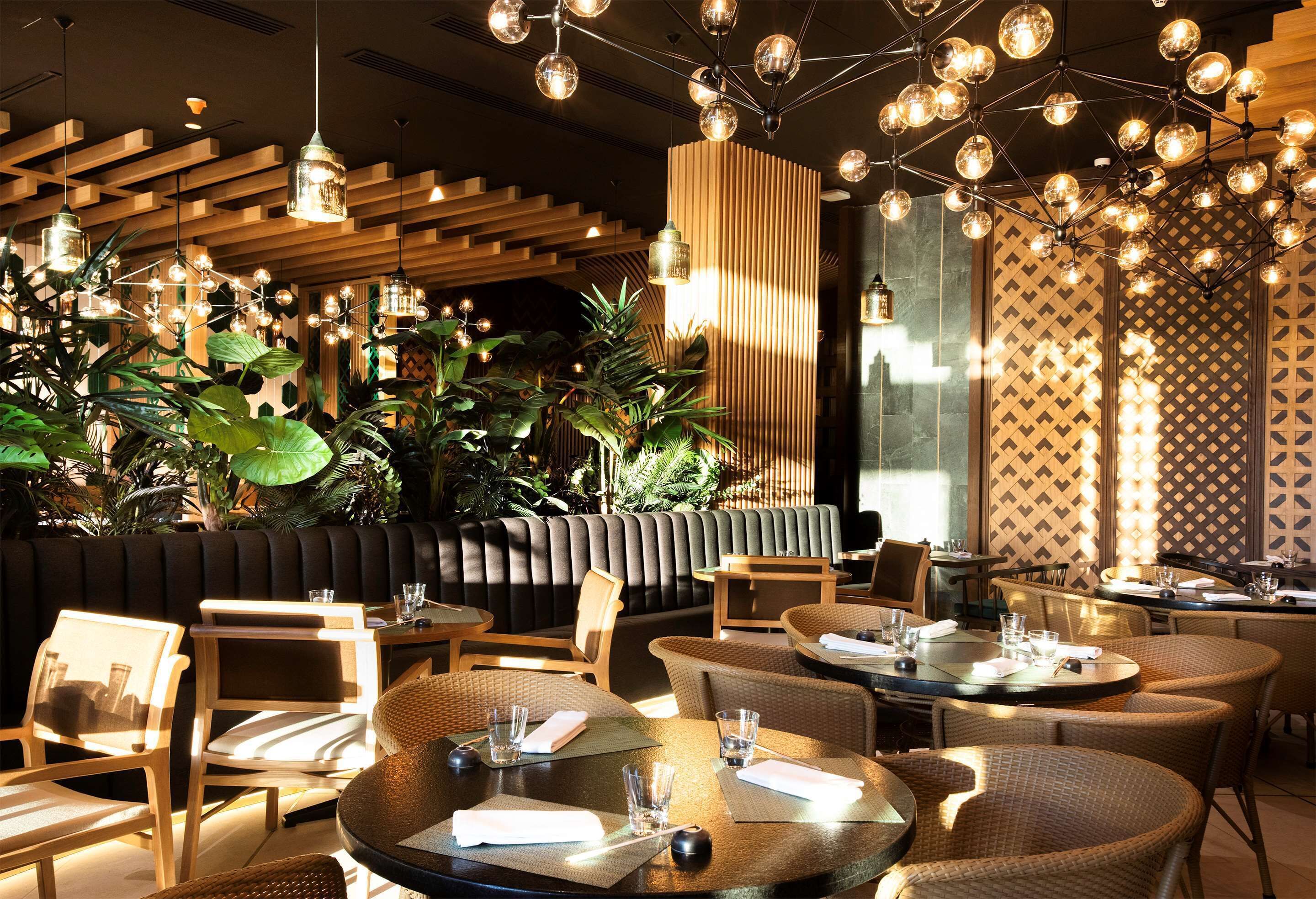 Een restaurant met ronde tafels, zachte stoelen, hangende lampen en binnenplanten.