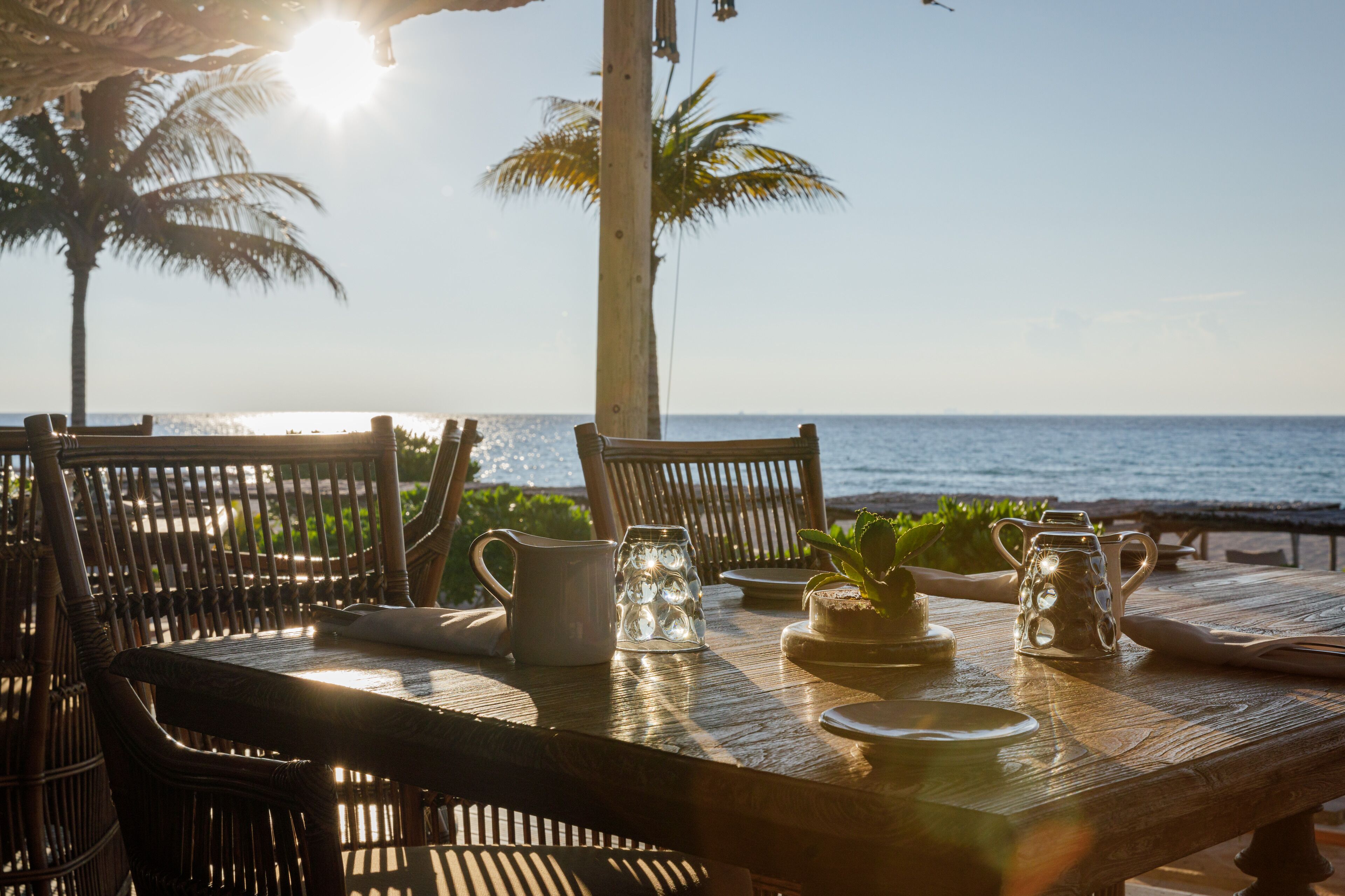 A wooden table set for dining with chairs, a pitcher, and a glass container, overlooking a beach with palm trees and the ocean.