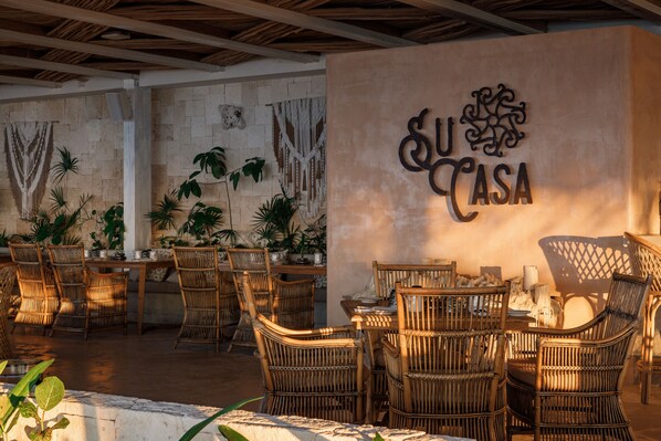 An outdoor dining area with wicker furniture and a wall with 'SU CASA' signage.