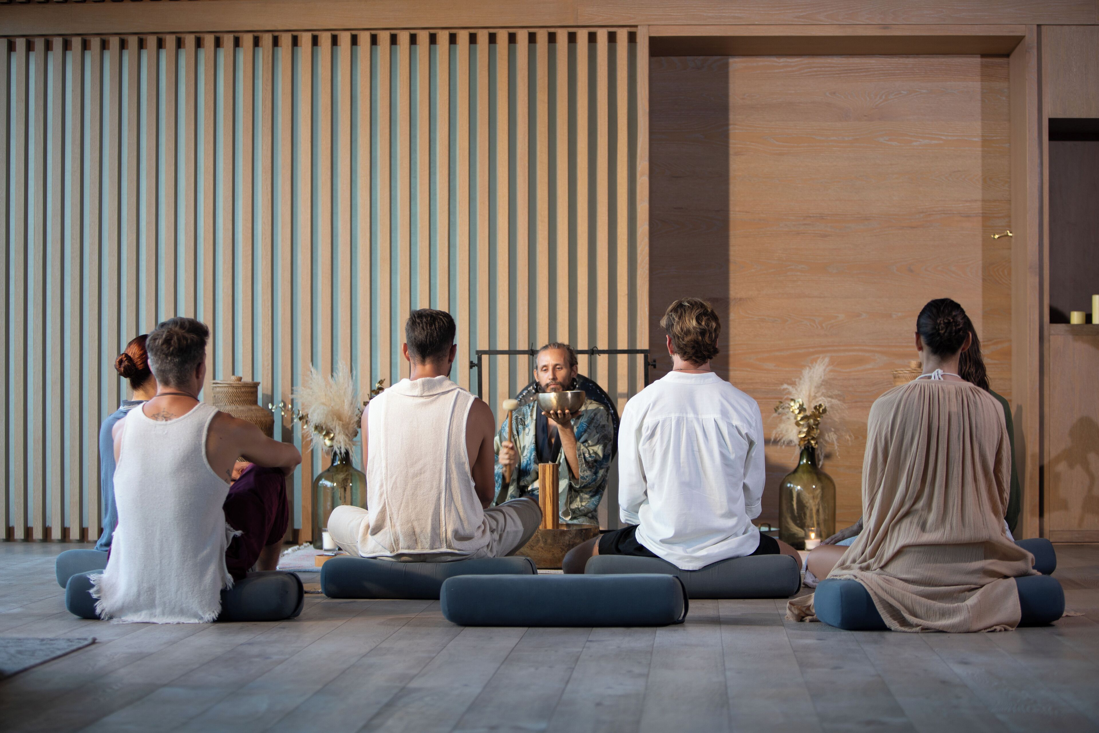 A group of people seated on cushions in a room with wooden slats on the wall and a wooden floor.