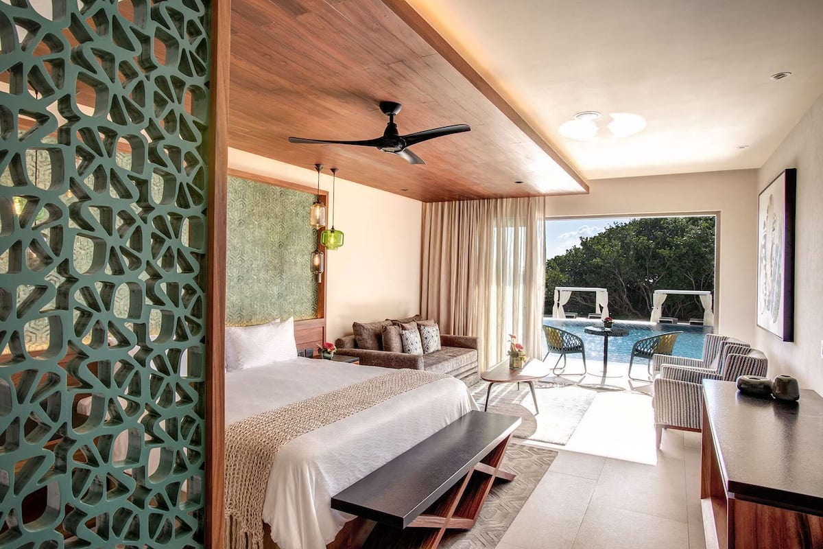 Habitación de hotel moderna con cama, sofá, mesa de comedor y vistas a una piscina al aire libre.
