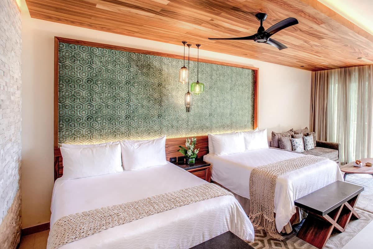Habitación de hotel con dos camas, techo de madera, cabecera estampada, ventilador de techo y una pequeña mesa.