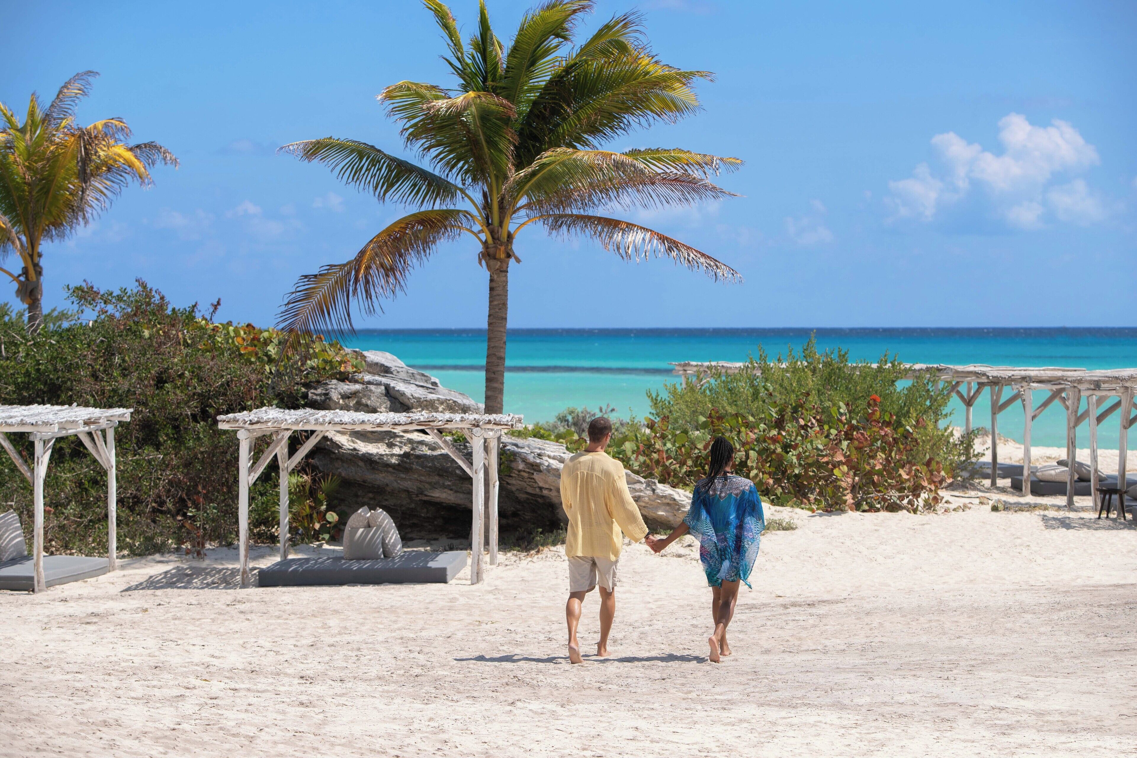 Una pareja caminando sobre una playa arenosa con cielo azul despejado y mar turquesa.