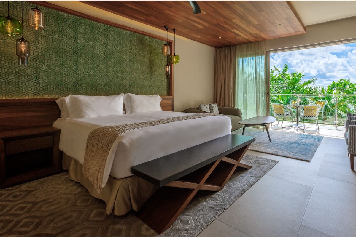 Habitación de hotel con cama, mesitas de noche, un banco, una pequeña mesa y un balcón con sillas.