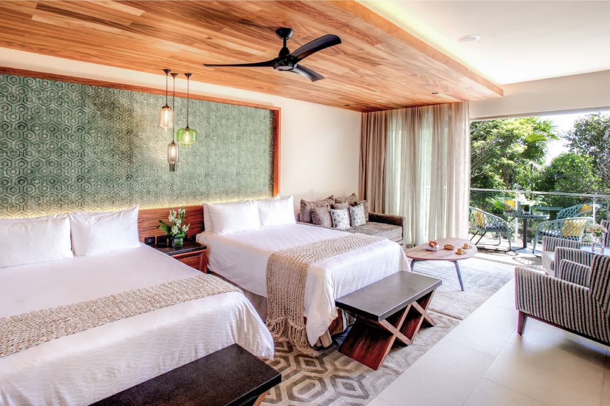 Habitación de hotel con dos camas, ventilador de techo, pared grande con diseño verde, balcón con mobiliario exterior y vistas a zonas verdes.