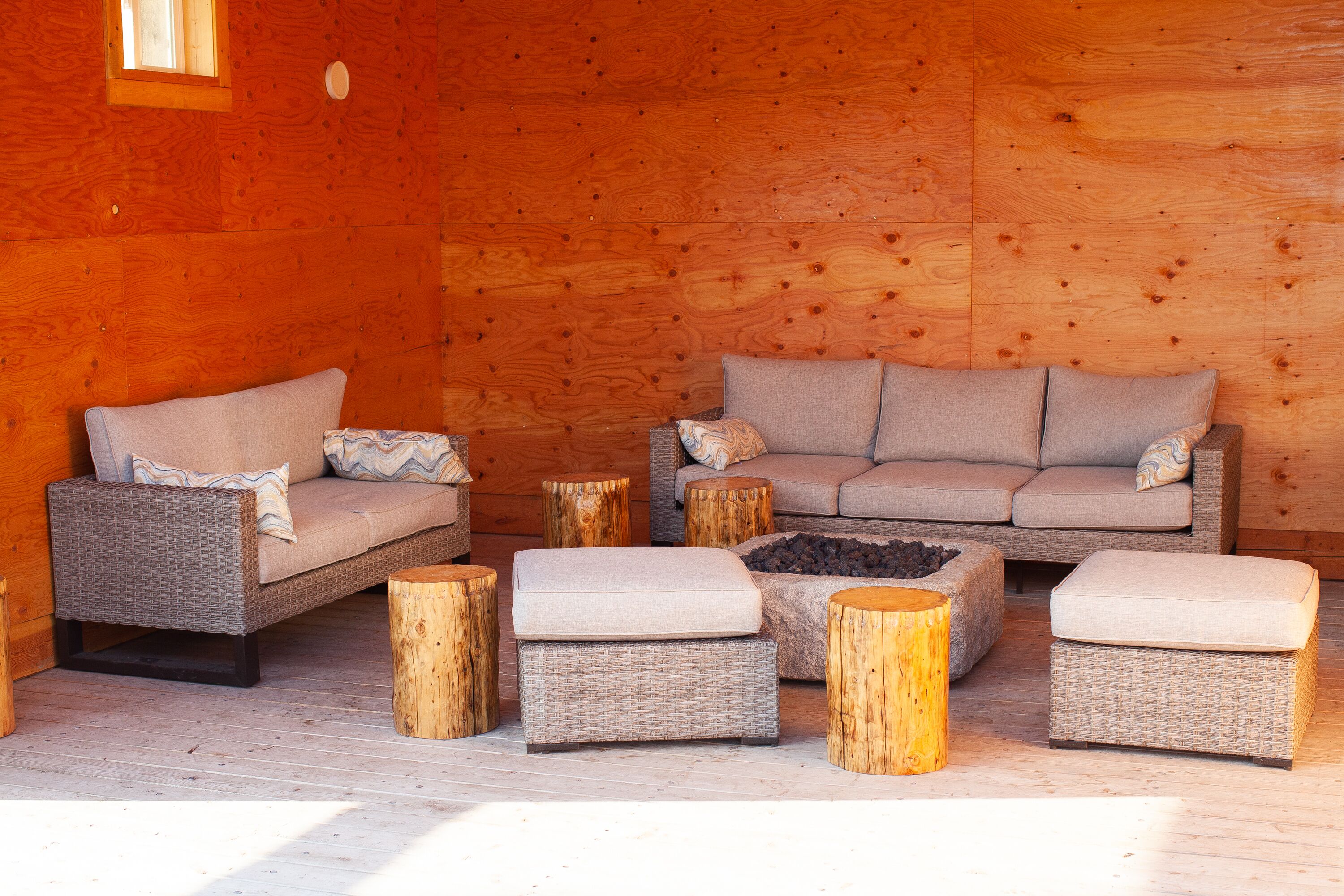 A cozy log cabin interior with wicker furniture, including a sofa and ottomans, and a central fire pit.