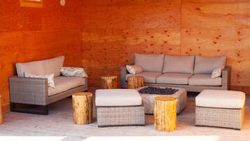A cozy log cabin interior with wicker furniture, including a sofa and ottomans, and a central fire pit.
