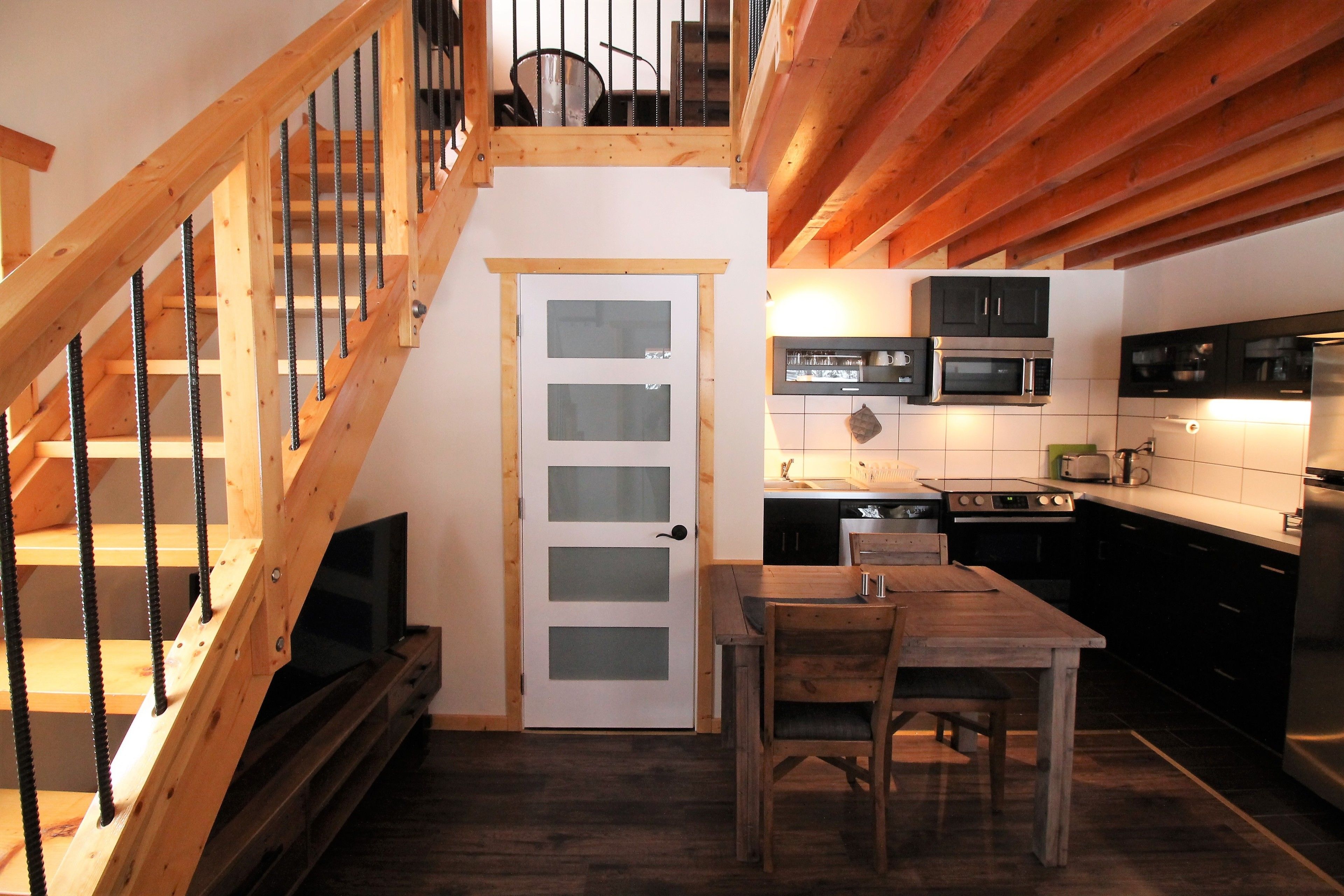 A wooden staircase leading to a loft bedroom with a bed and a TV. A kitchen with modern appliances and a dining area with a table and chairs is visible.
