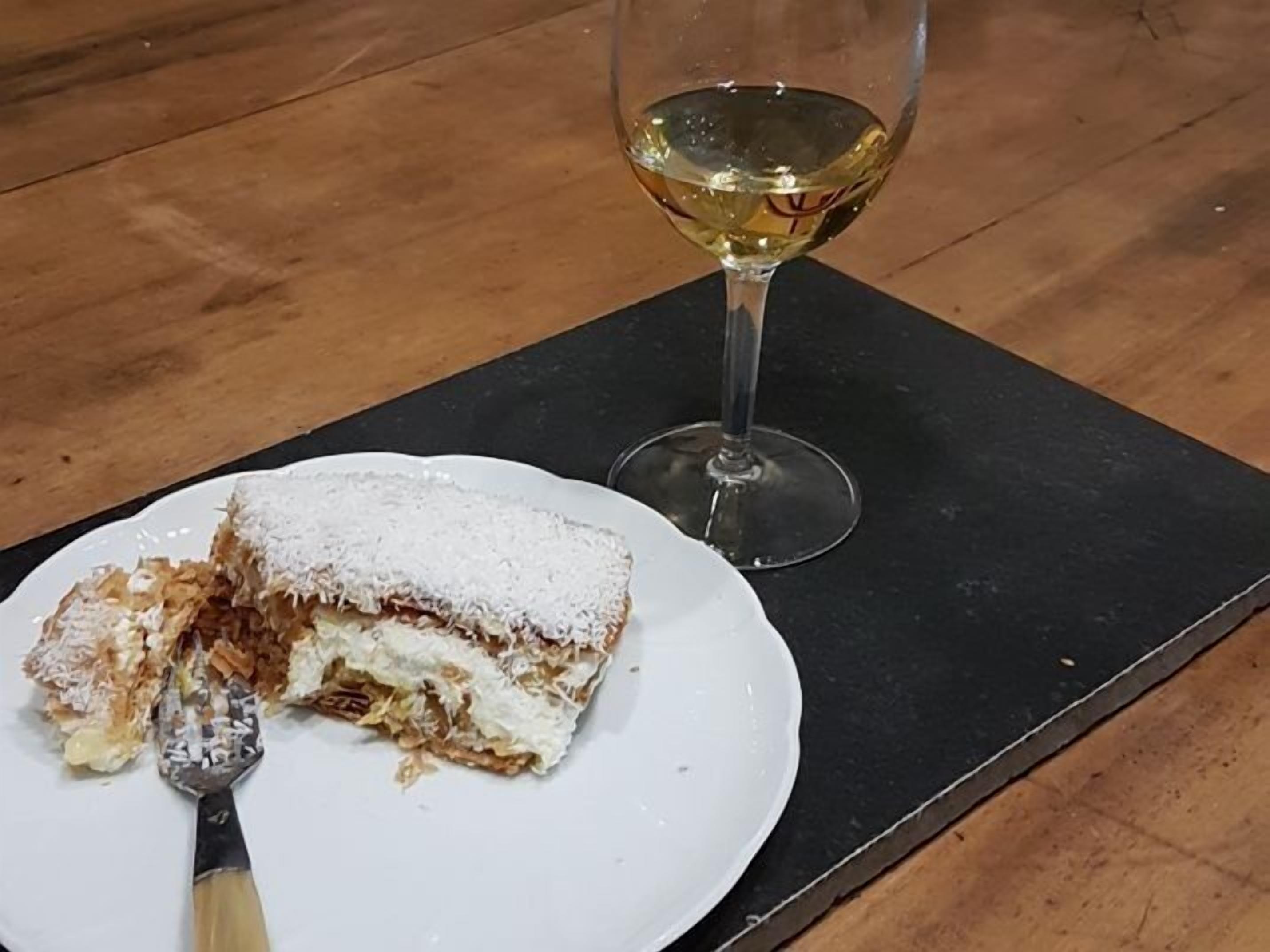 A slice of cake with powdered sugar on a white plate, a fork, and a glass of white wine on a black coaster.