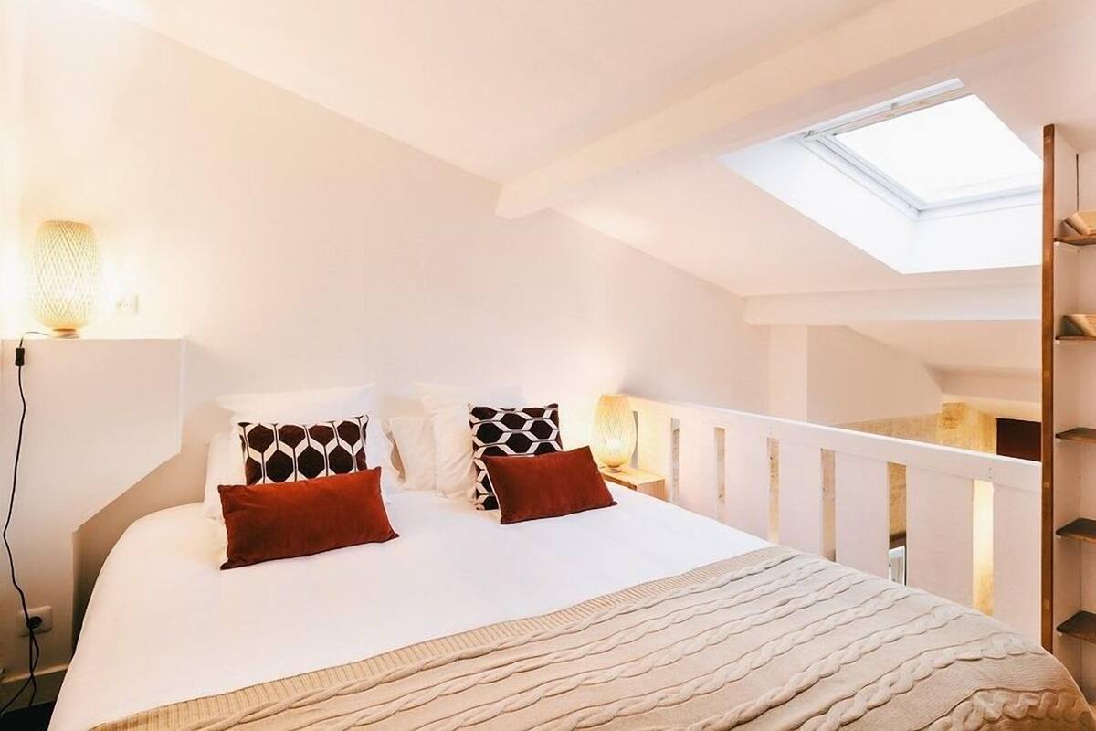 A bedroom with a skylight, a bed with patterned pillows, a bedside lamp, and a white railing.