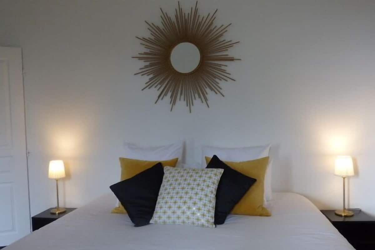 A neatly made bed with a white duvet, two bedside lamps, and a sunburst mirror above it.