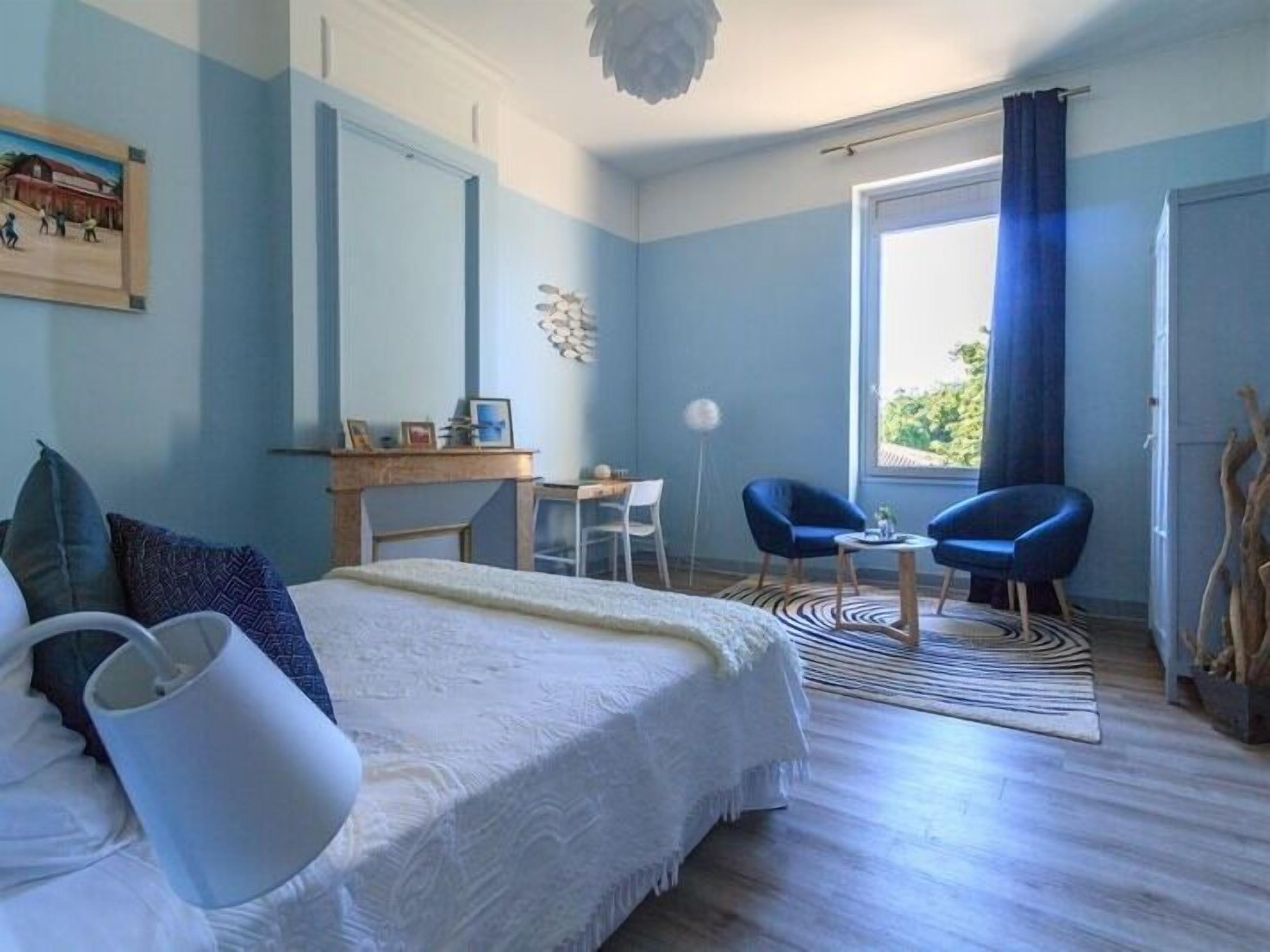 A bedroom with a bed, a desk, two chairs, a large window, and a view of greenery outside.