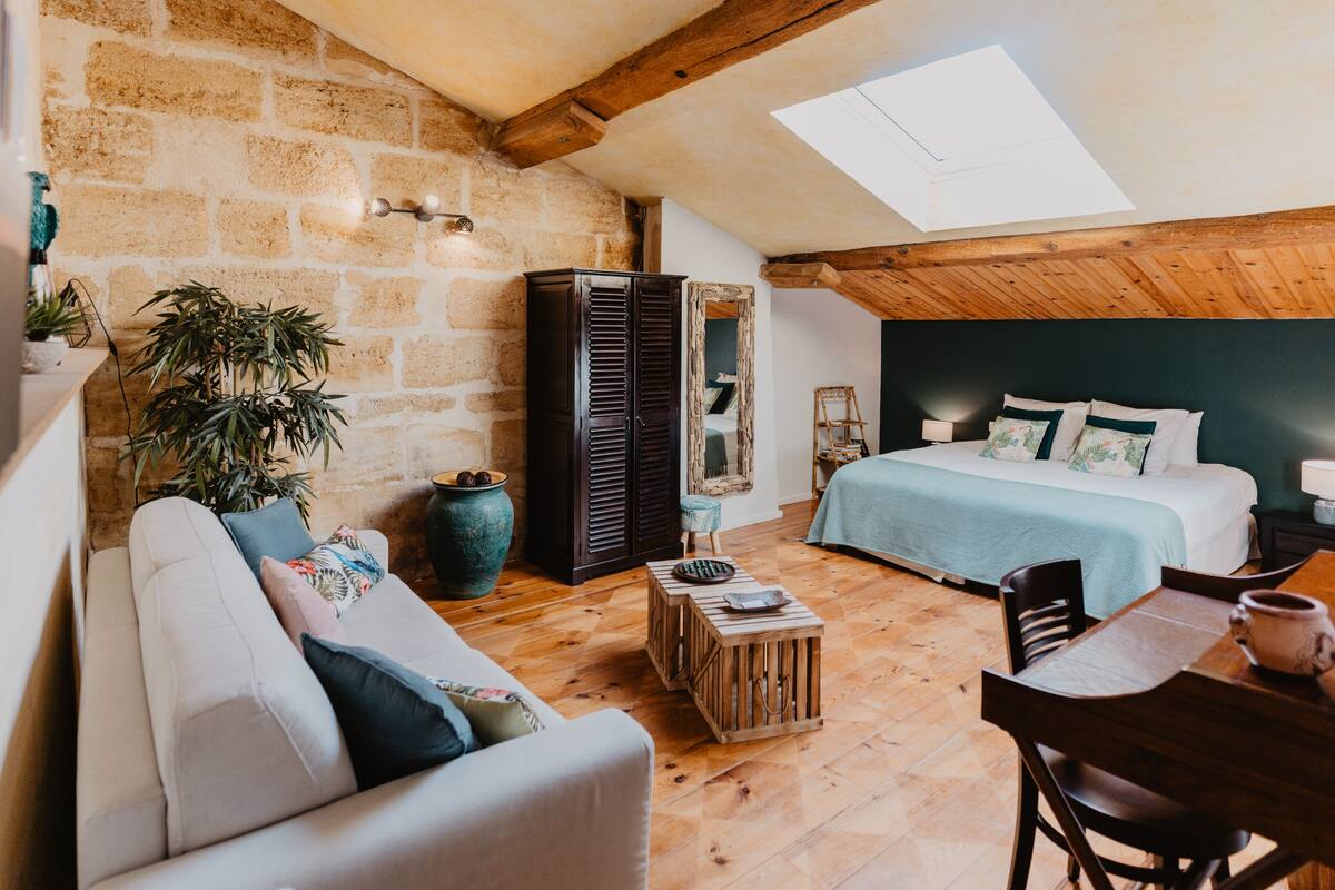 A cozy loft bedroom with a sloped ceiling, a large window, a bed with a blue bedspread, a wooden coffee table, and a sofa.