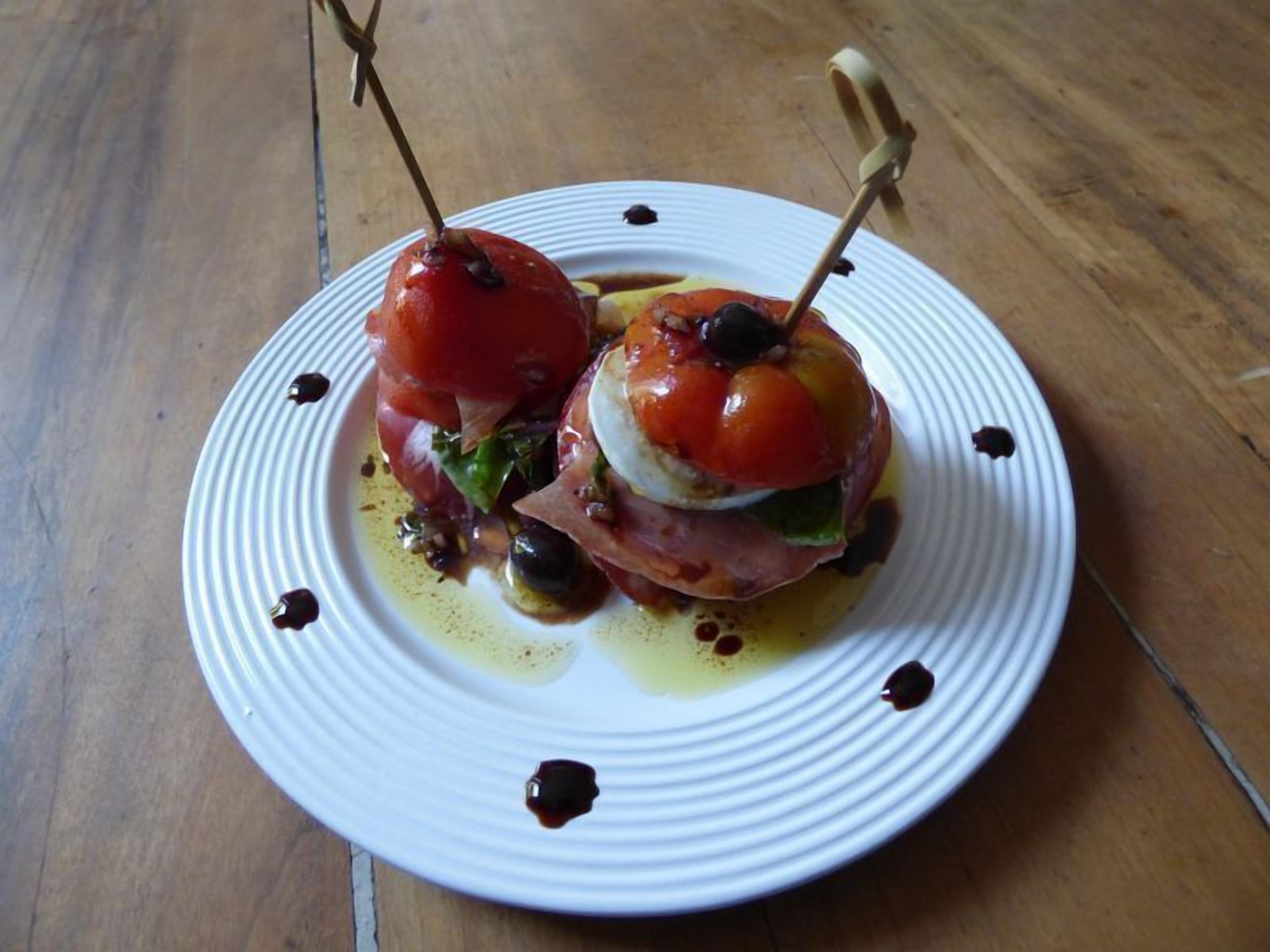 A plate with two stuffed tomatoes on skewers, olive oil, and balsamic glaze on a wooden table.