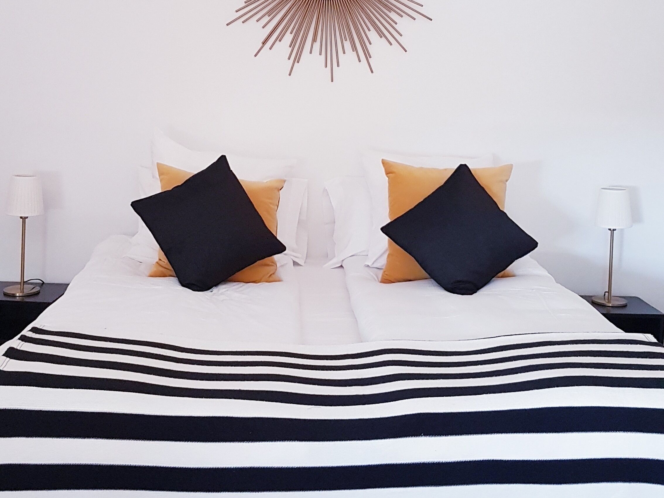 A neatly made bed with a striped bedspread, two black pillows, and two yellow pillows. There are two bedside tables with lamps on either side of the bed.