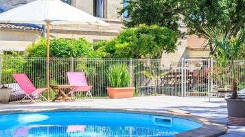 A swimming pool with a tiled edge, surrounded by a metal fence, and two lounge chairs under a white umbrella.