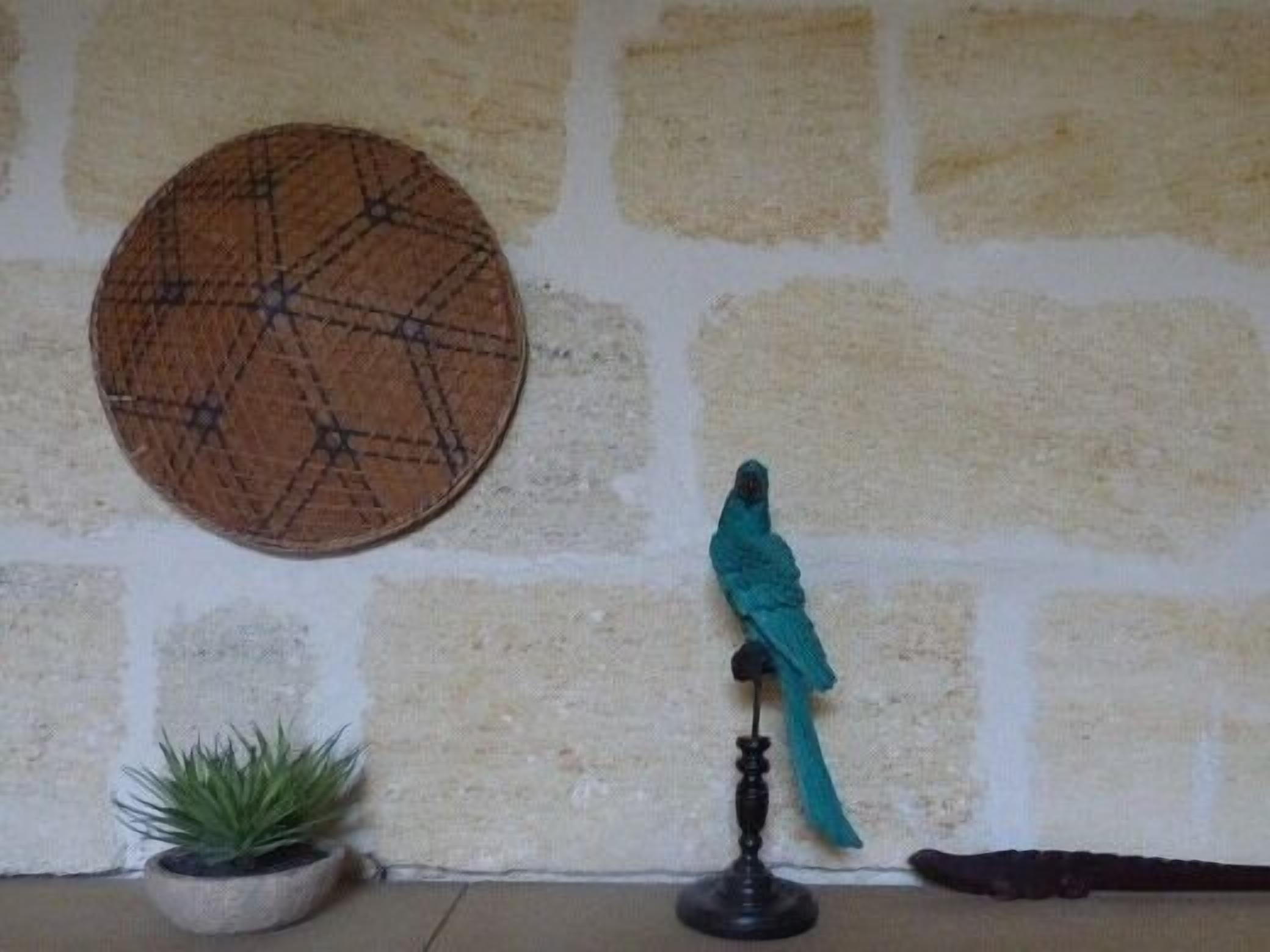 Een kamer met een stenen muur, een rond geweven mat, een kamerplant en een blauwgroene vogelbeeldje op een voetstuk.