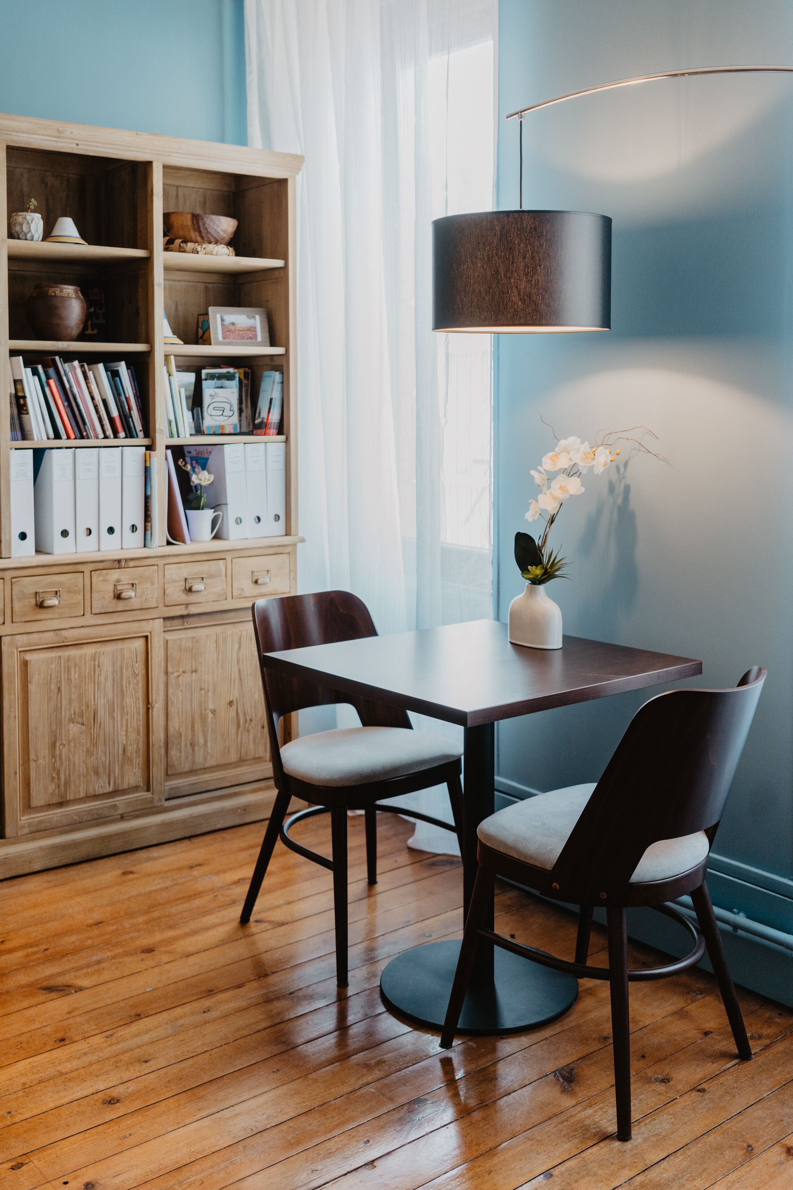 Un comedor con una mesa de madera, dos sillas, una estantería y una lámpara de pie.