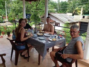 A group of people dining at an outdoor table with a checkered tablecloth, surrounded by greenery and a house in the background. - Villa ok (Unawatuna)