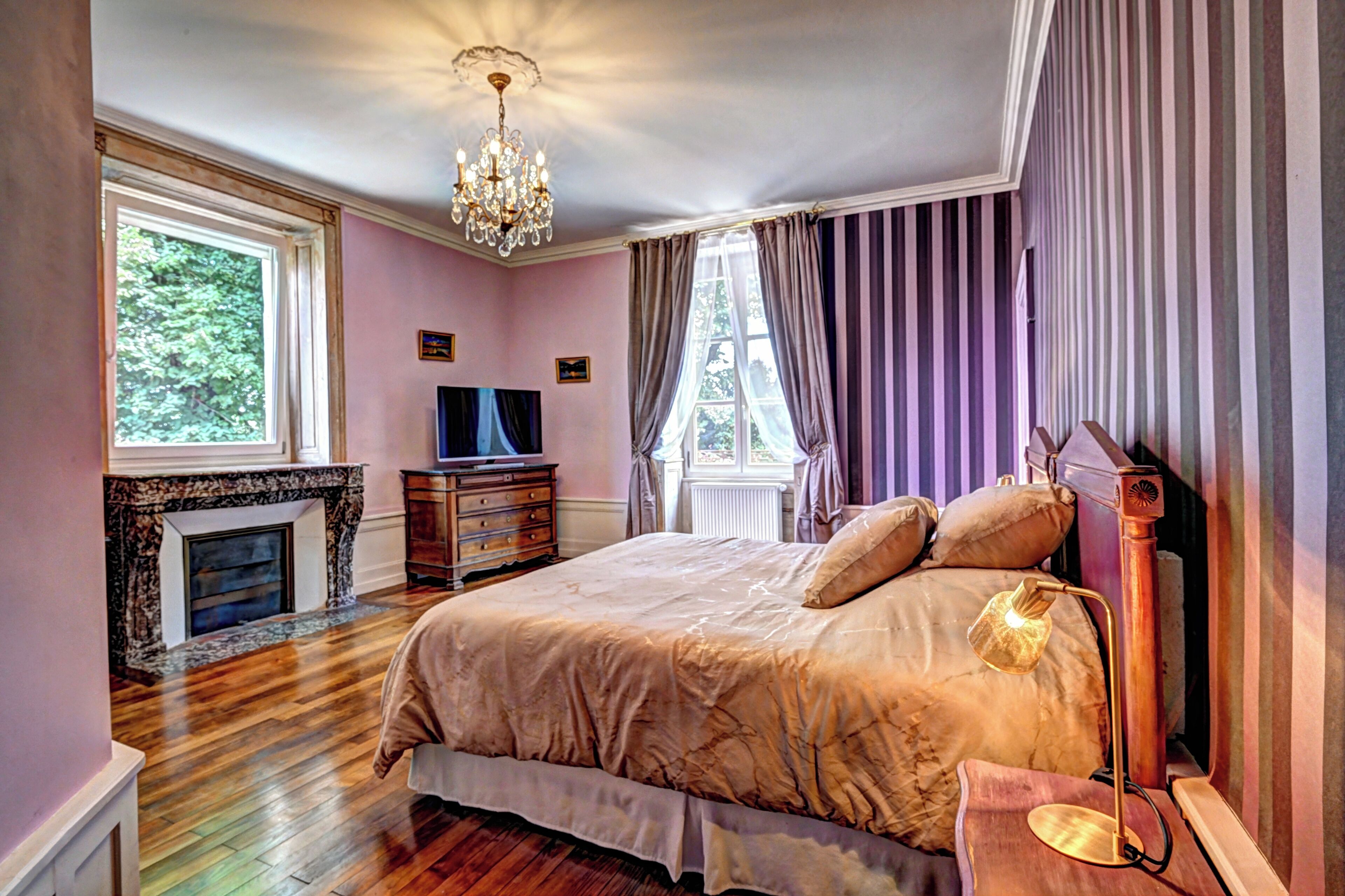 Un dormitorio con una cama grande, chimenea, candelabro y vista a árboles a través del ventanal.