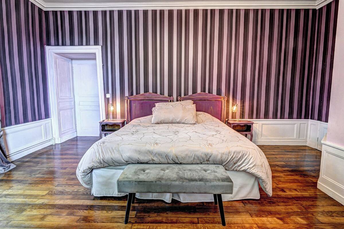 Une chambre avec un grand lit, un banc, deux tables de chevet avec des lampes et un mur à rayures.