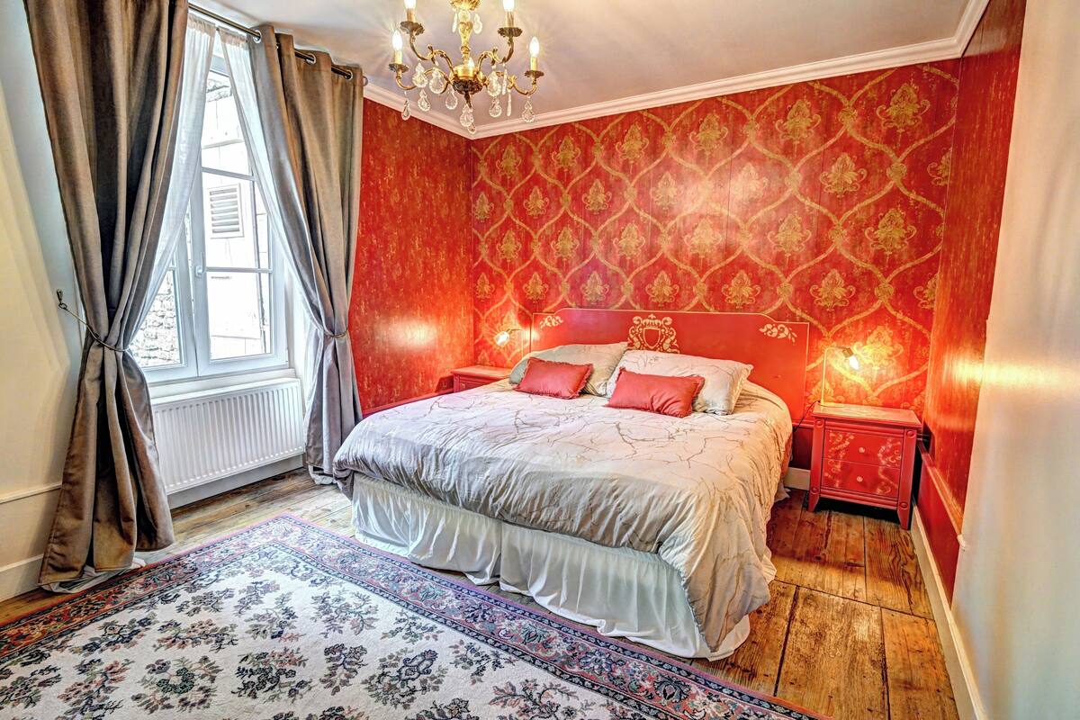 Une chambre à coucher avec un grand lit, un papier peint à motifs rouges, un lustre et une fenêtre avec des rideaux.