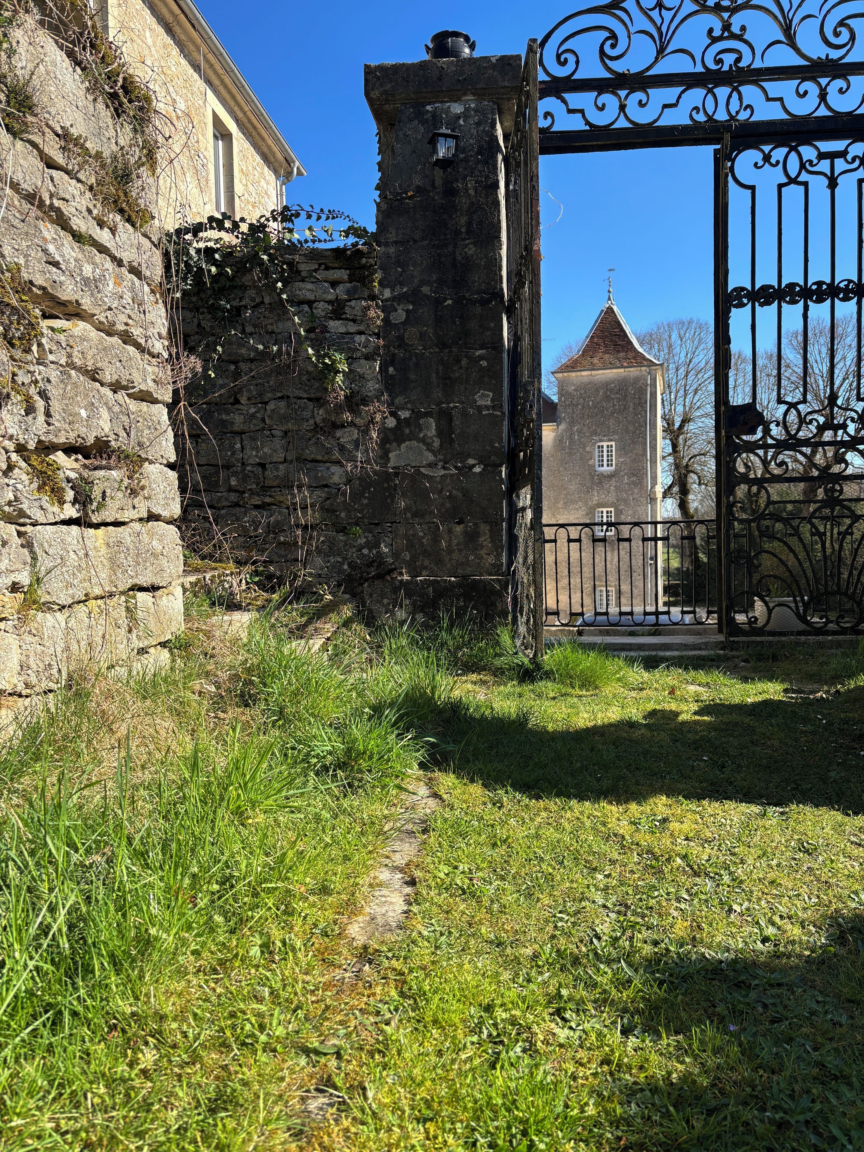 Un mur de pierre avec un portail en métal menant à un bâtiment doté d’un toit en tuiles.