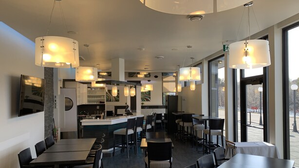 Ein modernes Restaurant mit hohen Tischen und Stühlen, Pendelleuchten und großen Fenstern mit Blick ins Freie.