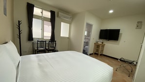 Quarto de hotel com uma cama grande, uma escrivaninha com duas cadeiras, uma televisão e um banheiro visível por uma porta aberta.