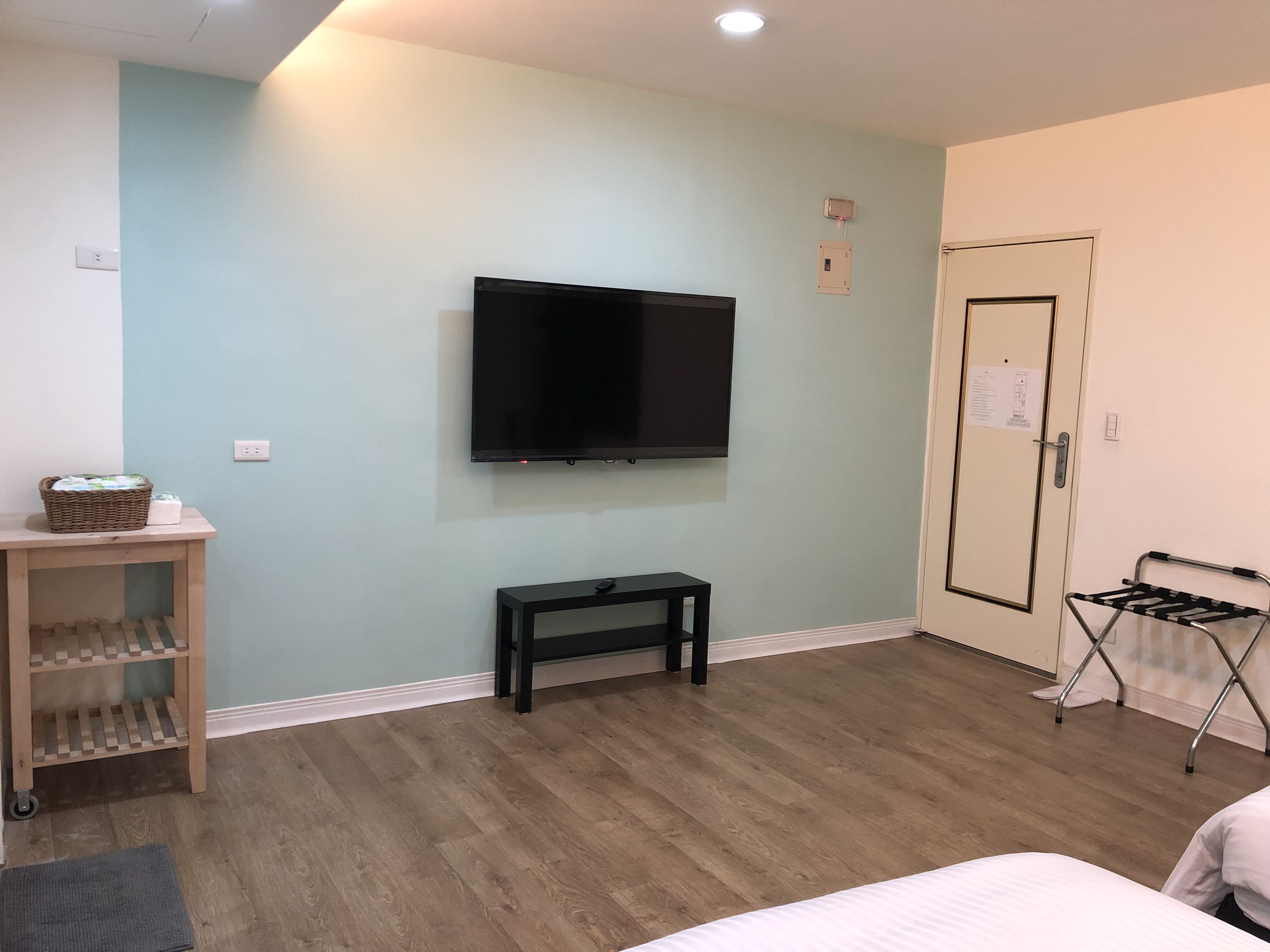 Een kamer met een wandgemonteerde flatscreen televisie, een klein houten tafeltje met een mand en flessen, een zwarte bank, een deur met een bericht en een opklapbare stoel.
