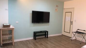 Een kamer met een wandgemonteerde flatscreen televisie, een klein houten tafeltje met een mand en flessen, een zwarte bank, een deur met een bericht en een opklapbare stoel.