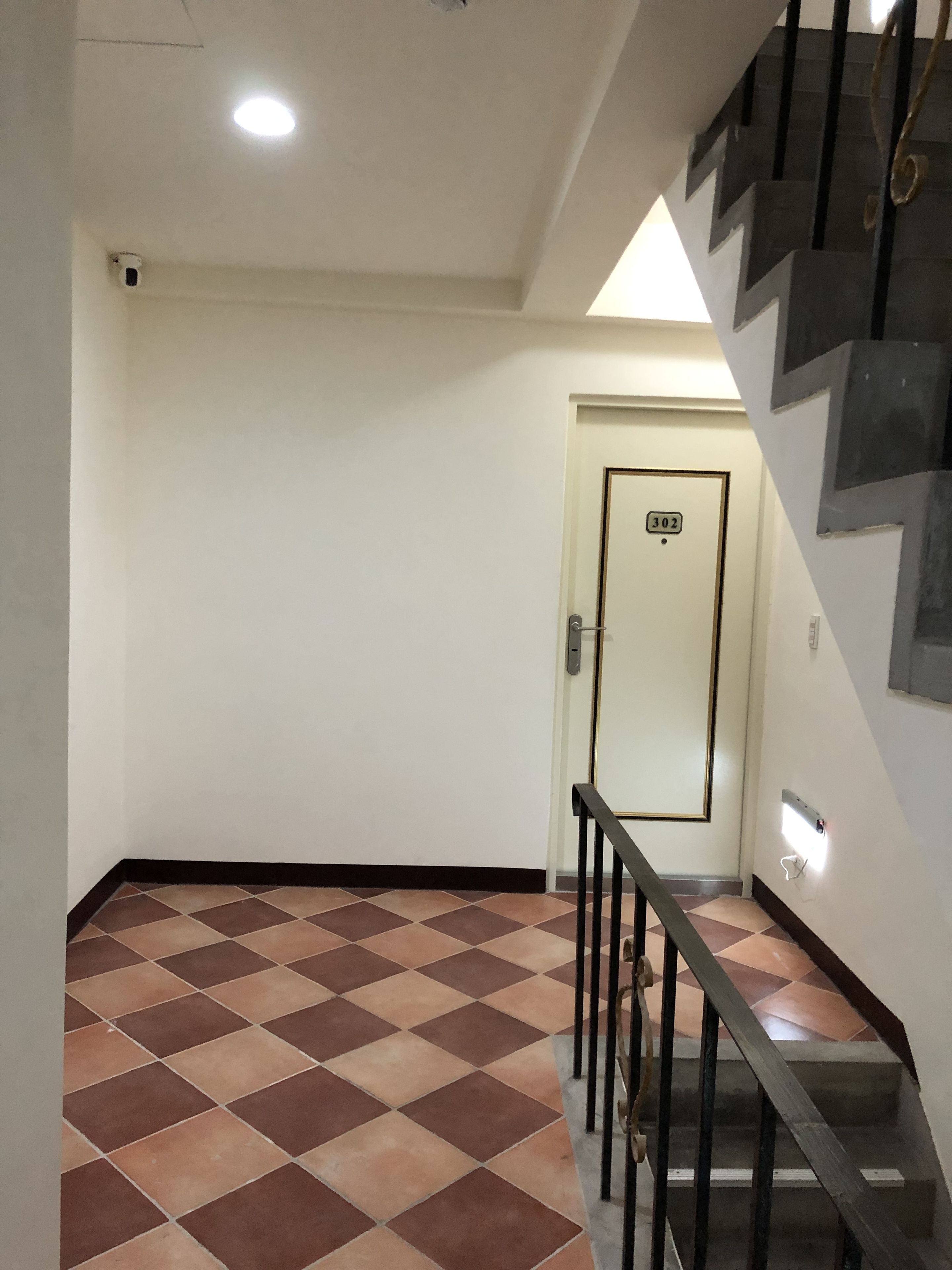 Un couloir avec un sol carrelé, une porte portant le numéro 302, un escalier avec des rampes noires et un luminaire au plafond.