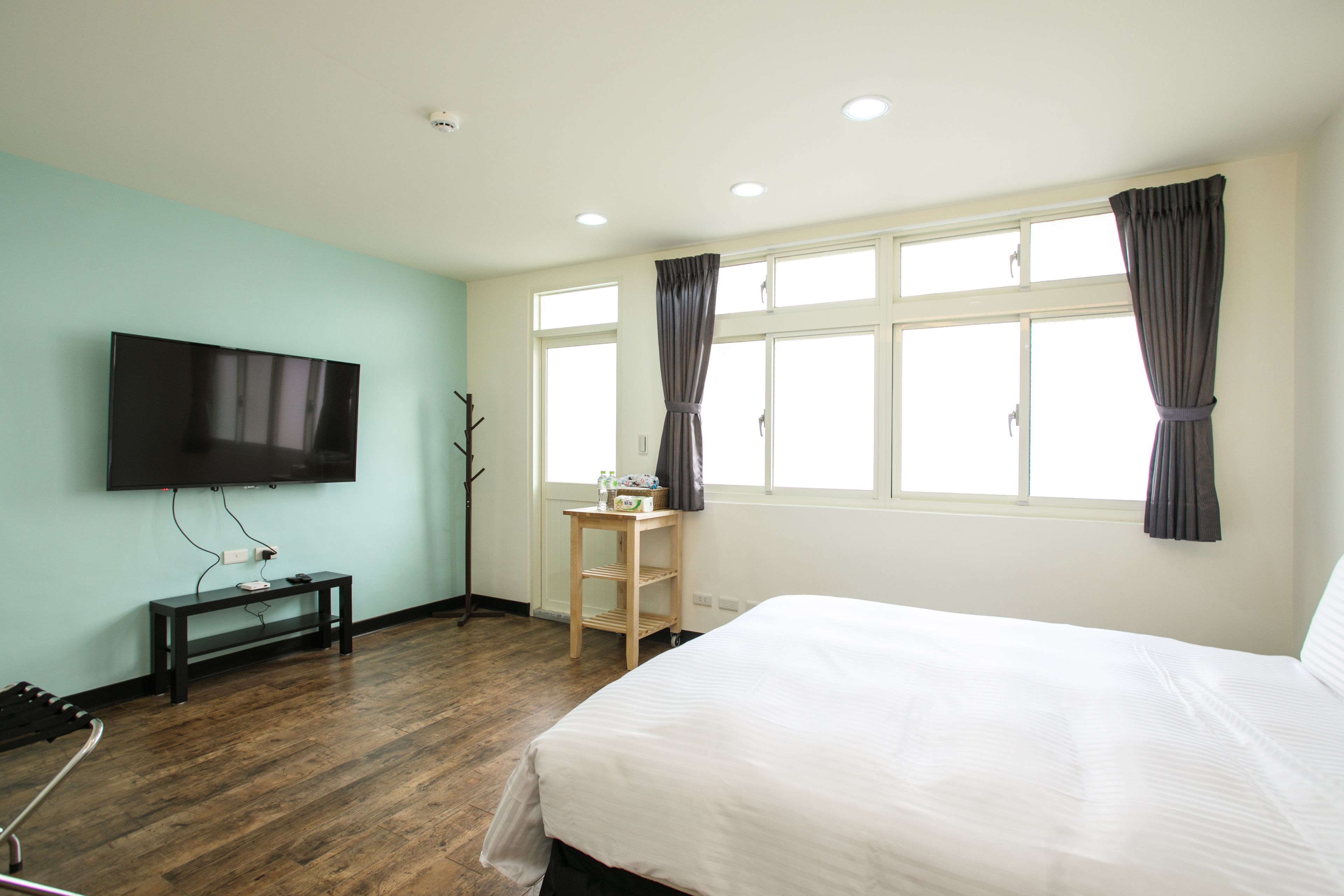 Habitación de hotel con cama, televisión montada en la pared, suelo de madera y ventana con cortinas.