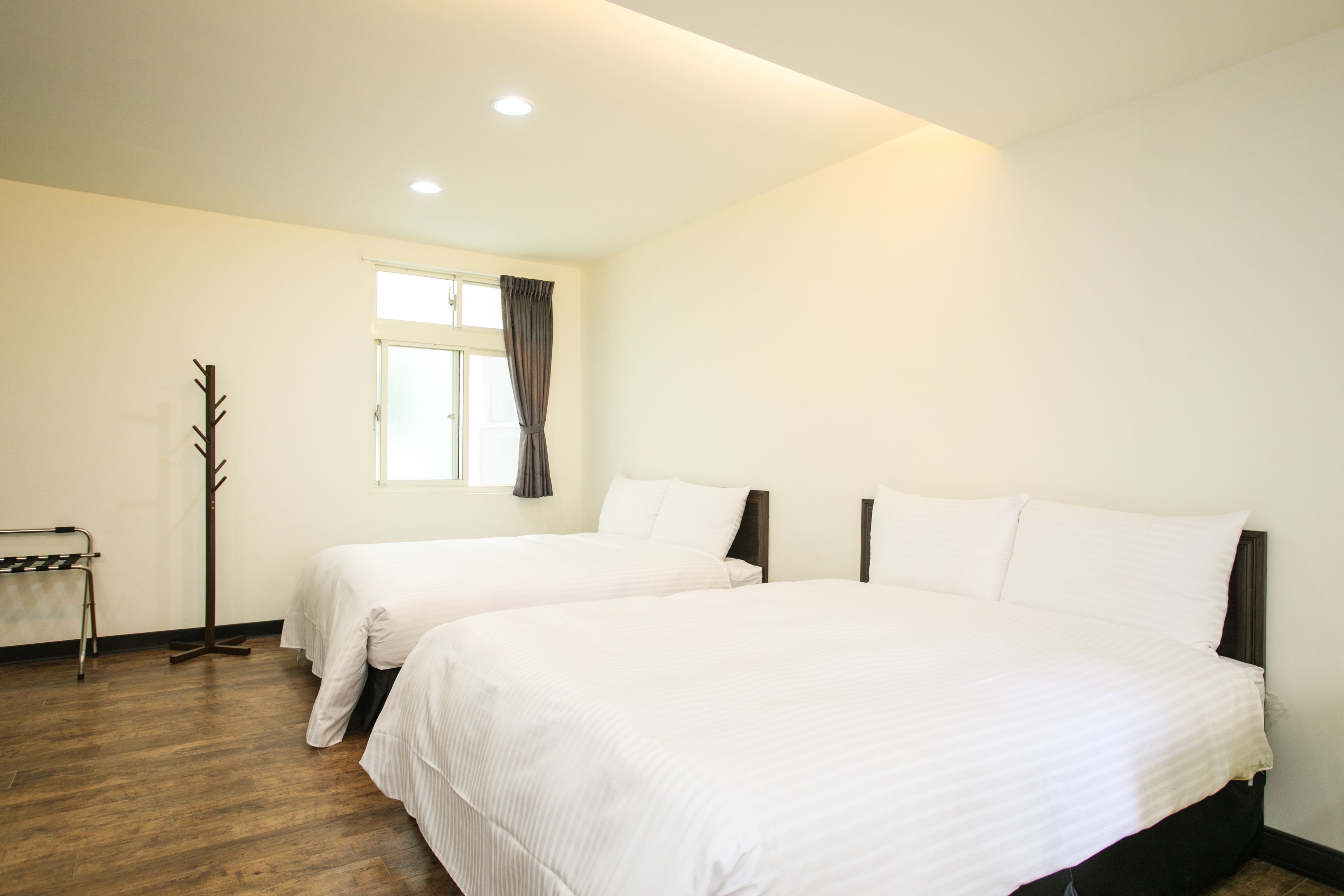 A hotel room with two beds, a coat rack, a window with curtains, and a wooden floor.